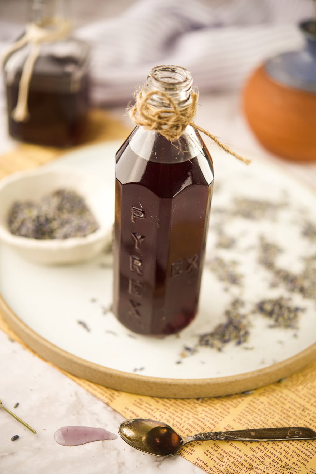 An open glass bottle of dark liquid, labeled PYREX and tied with twine, sits on a plate with scattered dried lavender buds, a spoon with some liquid, and a small bowl with more lavender nearby.