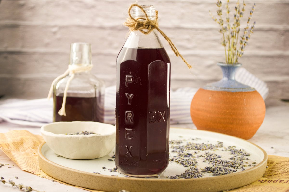 A bottle of Homemade Lavender Simple Syrup with a twine bow sits on a tray beside a white bowl of dried lavender, a second bottle, and a vase with lavender stems in a cozy, rustic setting.