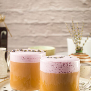 Two clear glass mugs filled with creamy lavender latte topped with dried lavender. The mugs sit on a round beige tray with a spoon, scattered lavender buds, and rustic decor in the background.