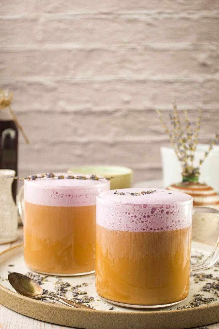 Two clear glass mugs filled with creamy lavender latte topped with dried lavender. The mugs sit on a round beige tray with a spoon, scattered lavender buds, and rustic decor in the background.