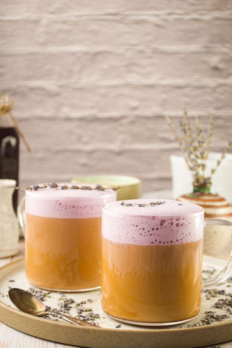 Two clear glass mugs filled with creamy lavender latte topped with dried lavender. The mugs sit on a round beige tray with a spoon, scattered lavender buds, and rustic decor in the background.
