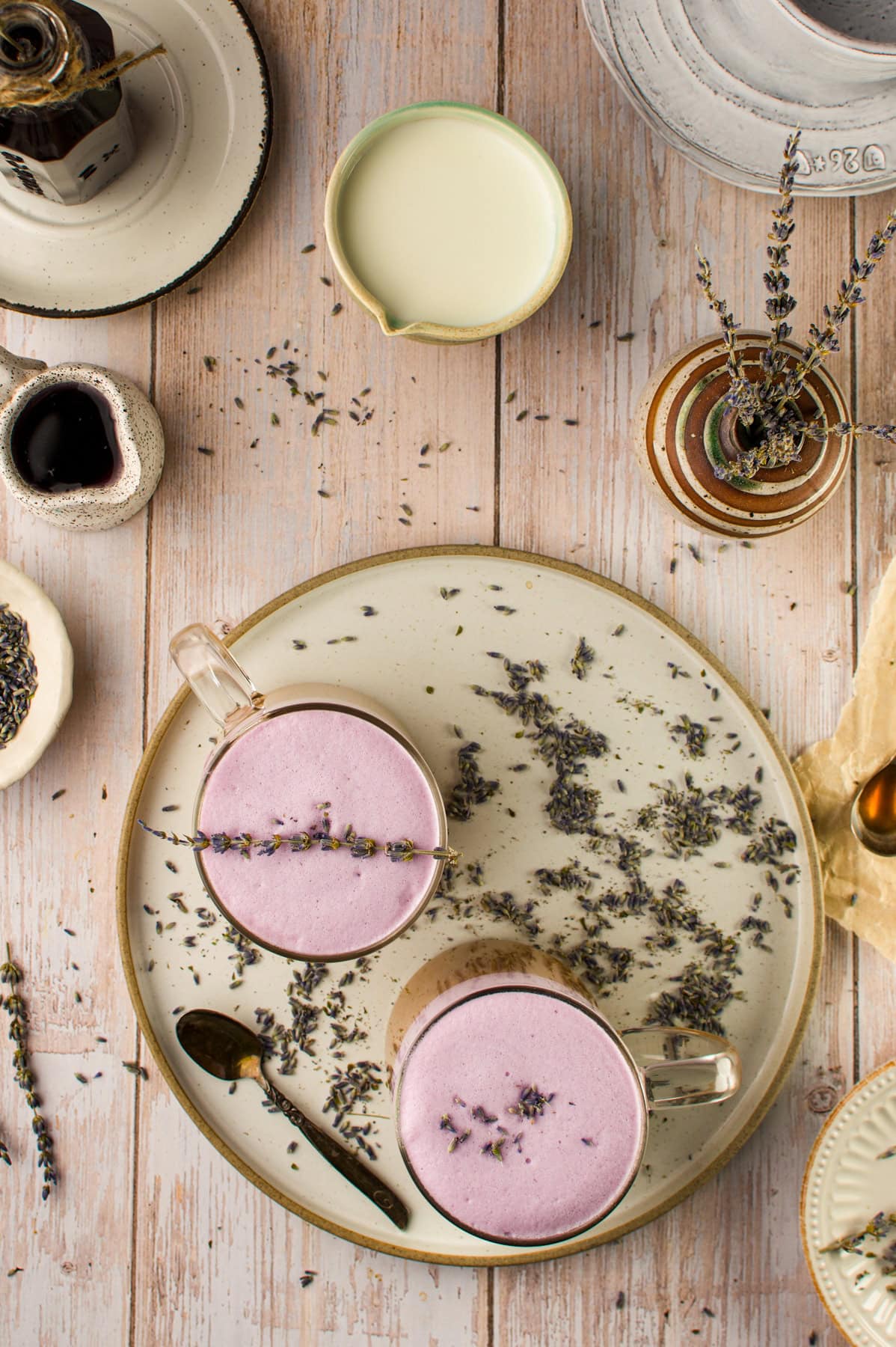 Two glass cups of lavender latte with frothy purple tops sit on a beige plate, surrounded by scattered dried lavender, a small pitcher of milk, a spoon, and ceramic bowls on a light wooden table.