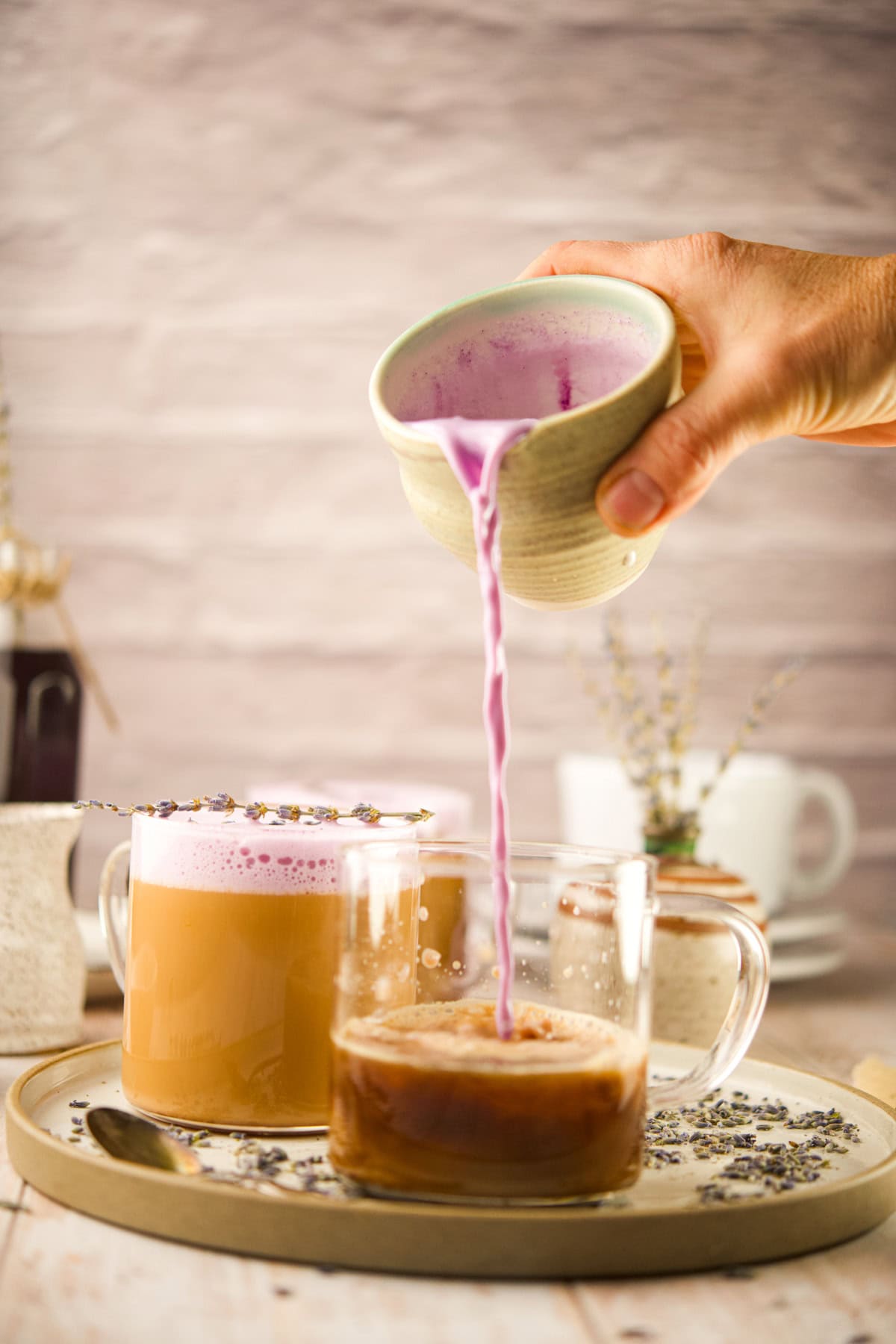 A hand pours a stream of  creamy lavender latte from a small ceramic pitcher into a glass mug of coffee on a wooden tray, surrounded by scattered lavender buds and another cup topped with foam and lavender.