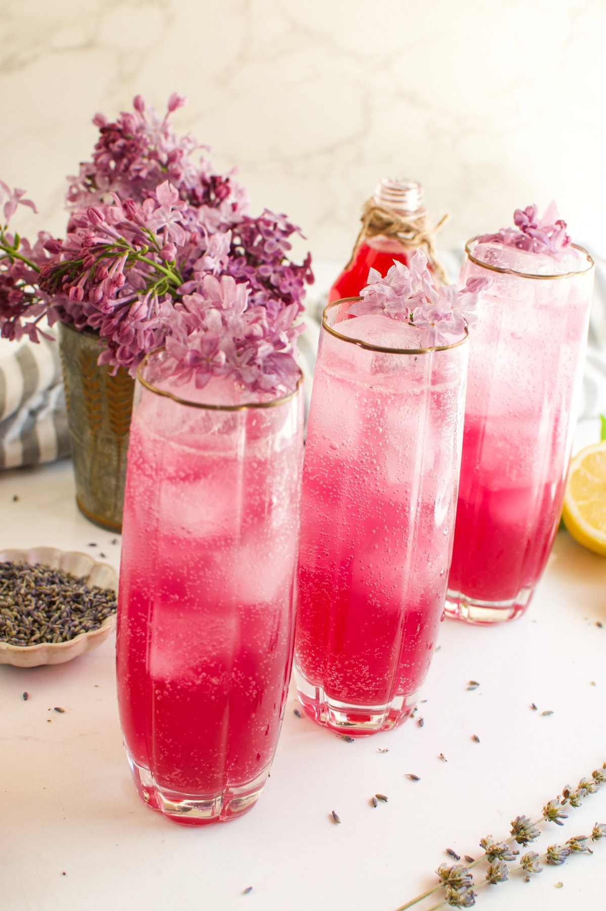 Three tall glasses filled with pink sparkling beverages, garnished with purple lilac flowers and ice, are placed on a white surface with scattered lilacs, lemon, and a bottle of pink drink in the background.