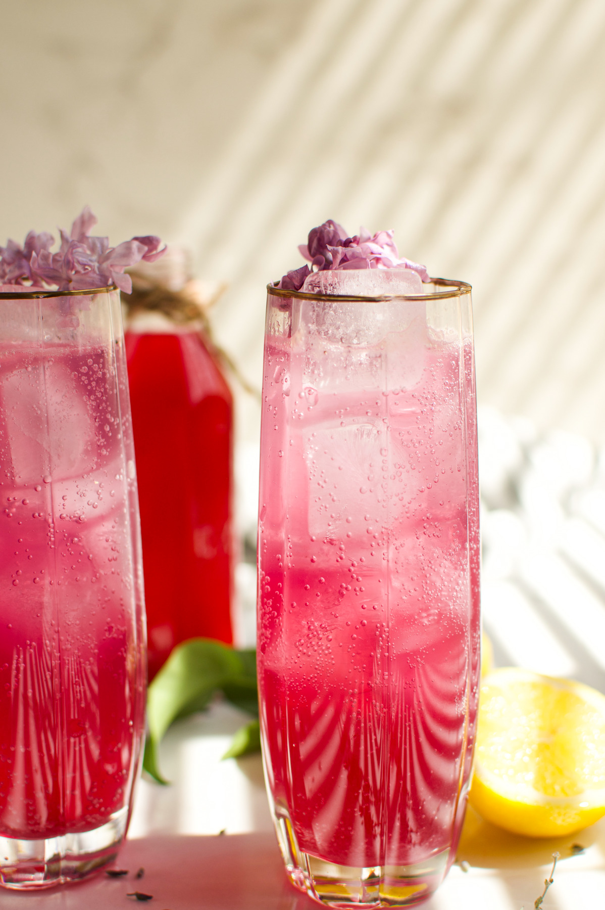 Two tall glasses filled with a Lilac Lavender Spritzer topped with purple flowers, sitting on a white surface. A lemon wedge and a red bottle are in the background with sunlight casting shadows.