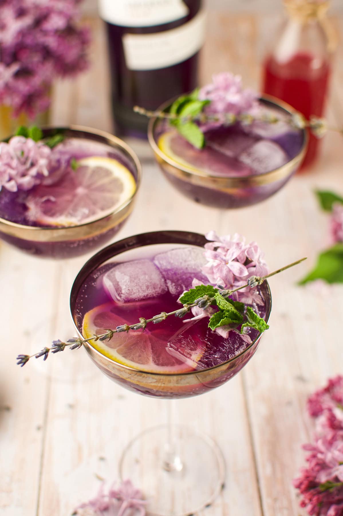 A close-up of purple cocktails in coupe glasses, garnished with ice cubes, lemon slices, mint, and small purple flowers, set on a light wooden surface with more flowers and a bottle in the background.