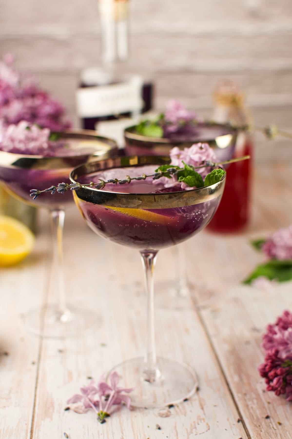 Three elegant cocktail glasses filled with a deep purple drink, garnished with lavender sprigs, mint, and orange slices. Lilac flowers and a lemon are scattered on a rustic wooden surface, with bottles blurred in the background.
