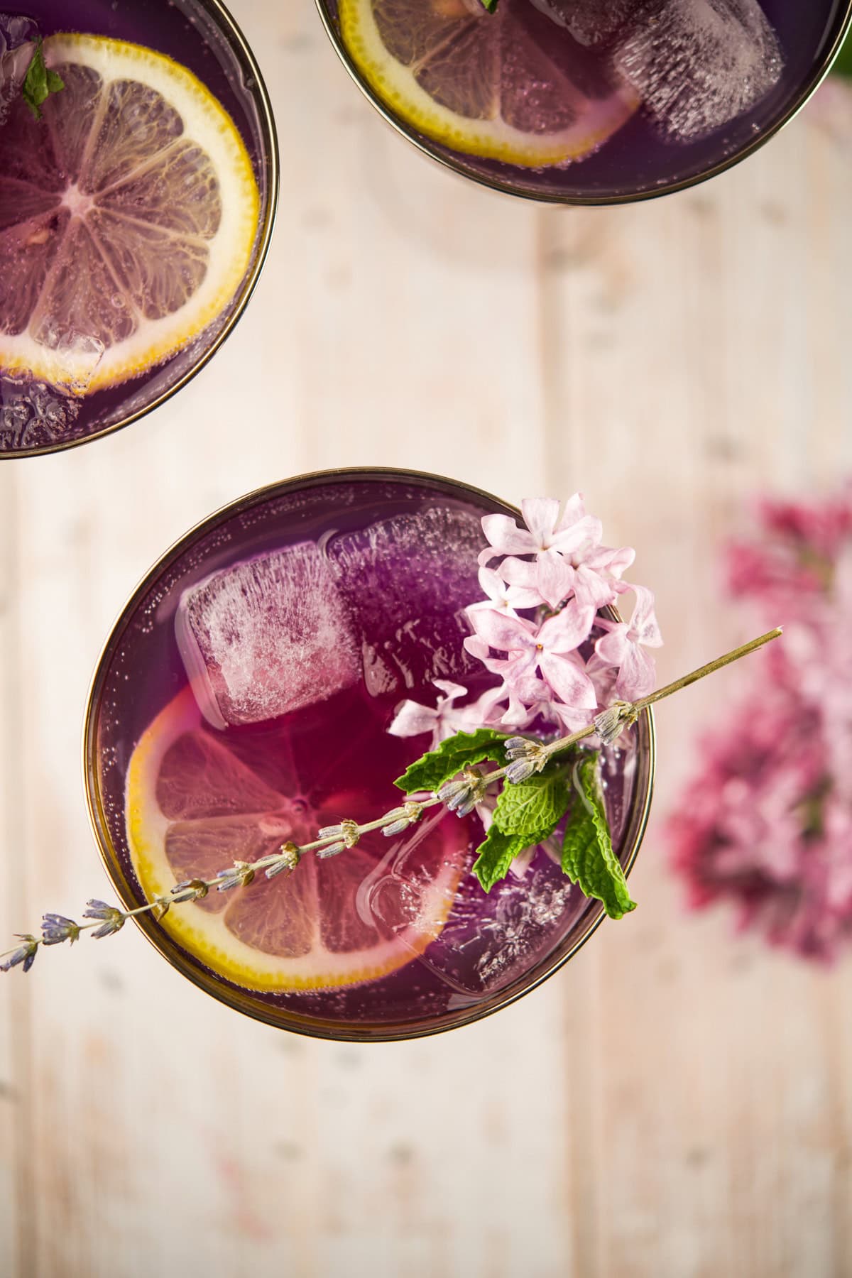 Overhead view of a purple drink with ice cubes, lemon slices, and a garnish of lavender flowers and mint in a glass, set on a light wooden surface with blurred flowers in the background.