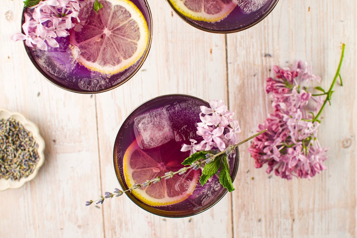 Overhead view of purple cocktail drinks with ice cubes, lemon slices, and lilac flowers, garnished with mint and lavender on a rustic white wooden table. A small dish of dried lavender is nearby.