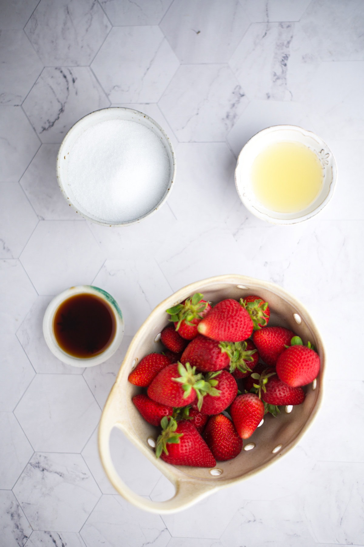 Overhead view of fresh strawberries in a colander, with bowls containing sugar, vanilla extract, and lemon juice on a white hexagon-tiled surface.