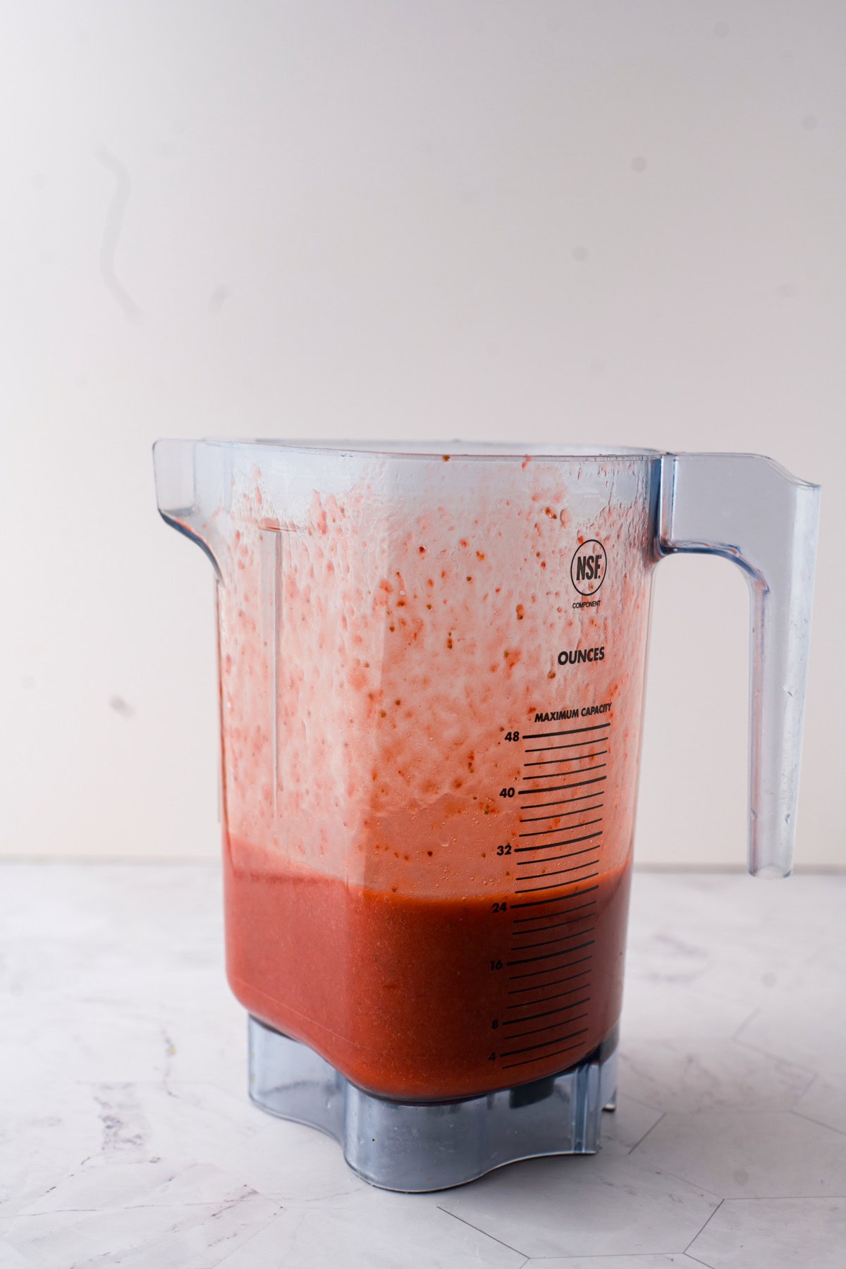 A blender pitcher filled with a red, frothy liquid such as a fruit smoothie or sauce, sitting on a light-colored, marble surface. The pitcher has measurement markings on the side.