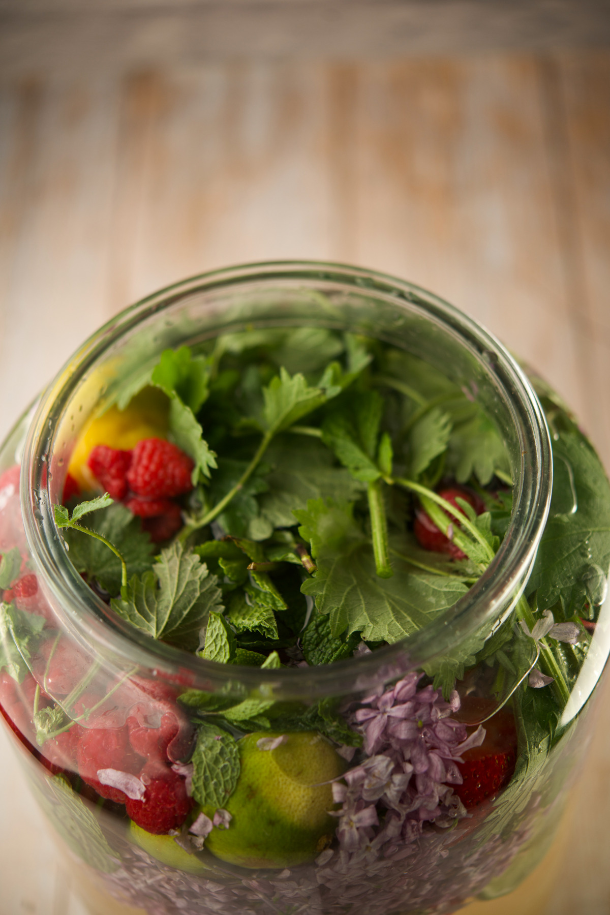 A glass jar filled with fresh herbs, leafy greens, raspberries, lime slices, and purple flower petals, set on a wooden surface.