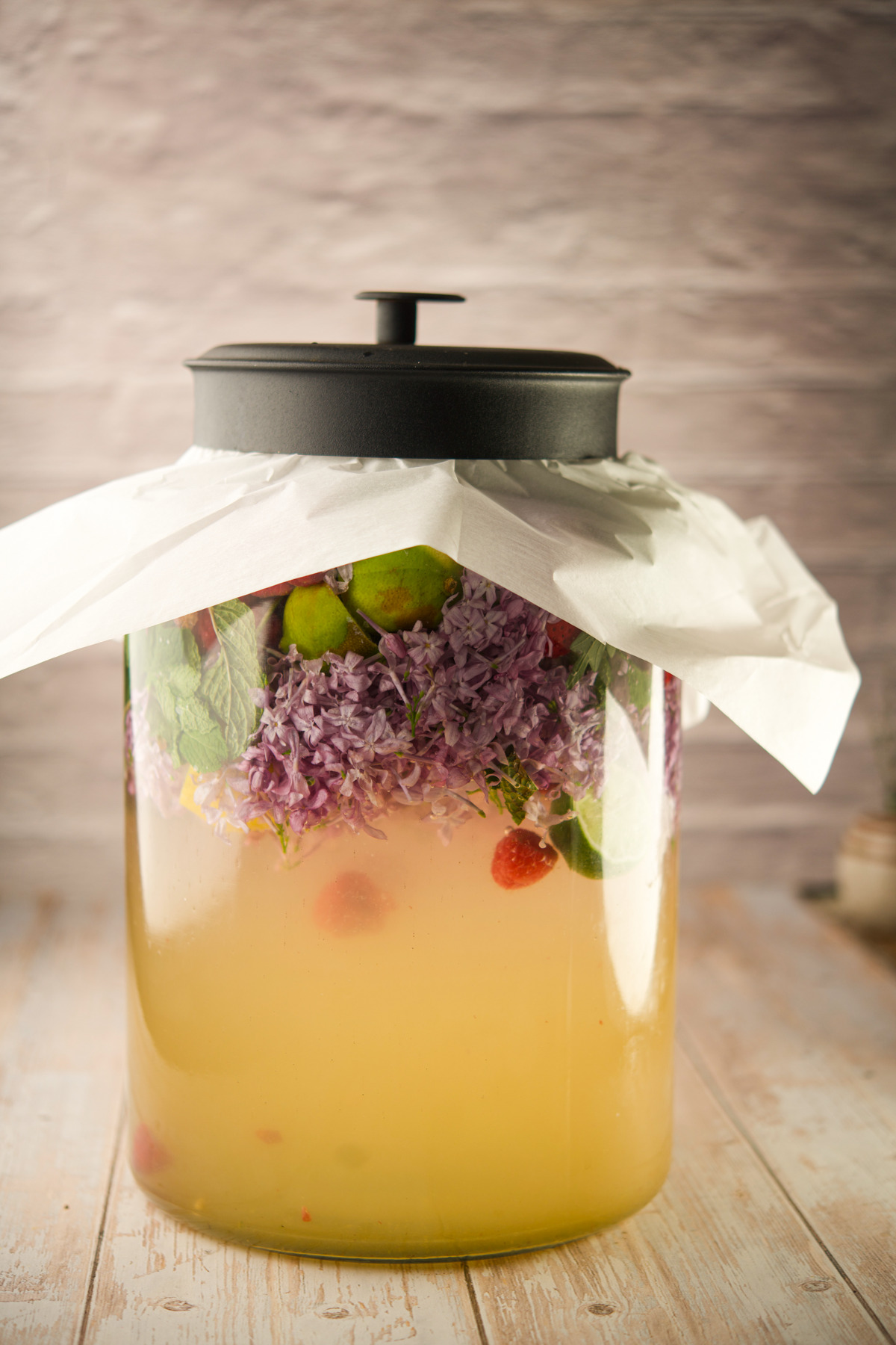 A large glass jar filled with a yellowish liquid, floating lilacs, limes, mint leaves, and berries, covered with a white cloth and topped with a black lid, sits on a light wooden surface.