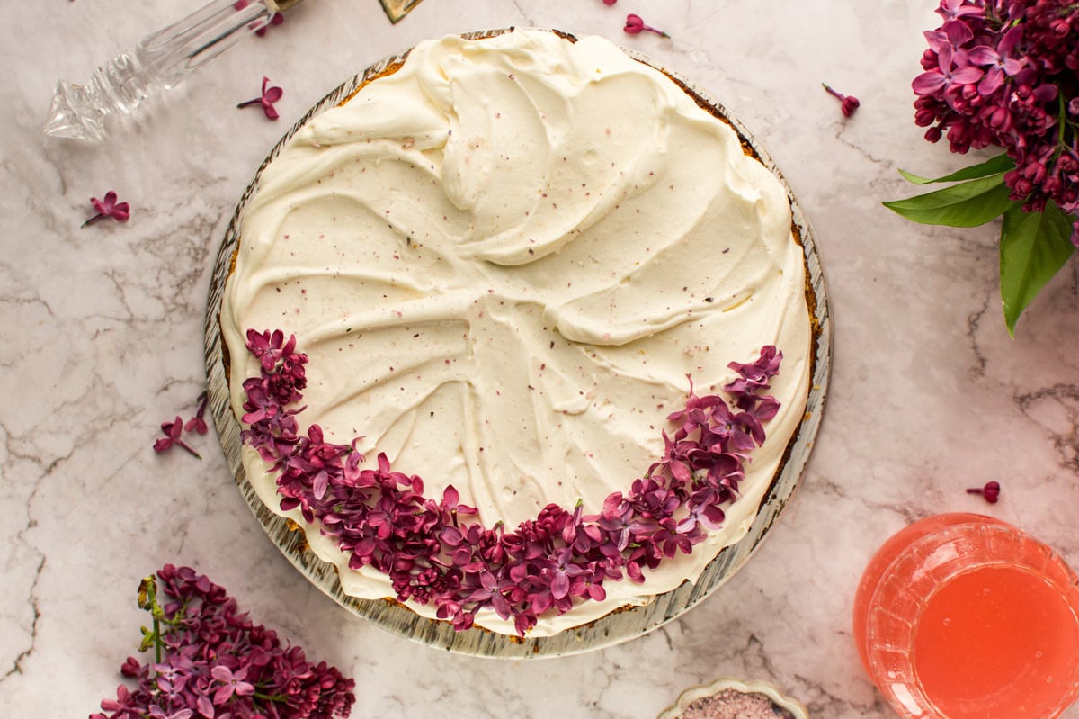 A pie topped with swirled white frosting and a crescent of purple lilac flowers, placed on a marble surface with clusters of lilacs and a pink drink nearby.
