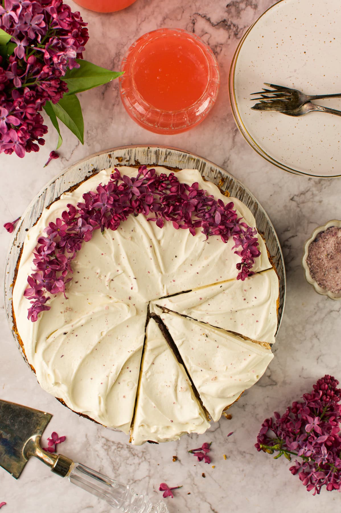A Lilac Cheesecake with purple lilac flowers, two slices cut, surrounded by plates, forks, a drink, and lilac sprigs on a marble surface.
