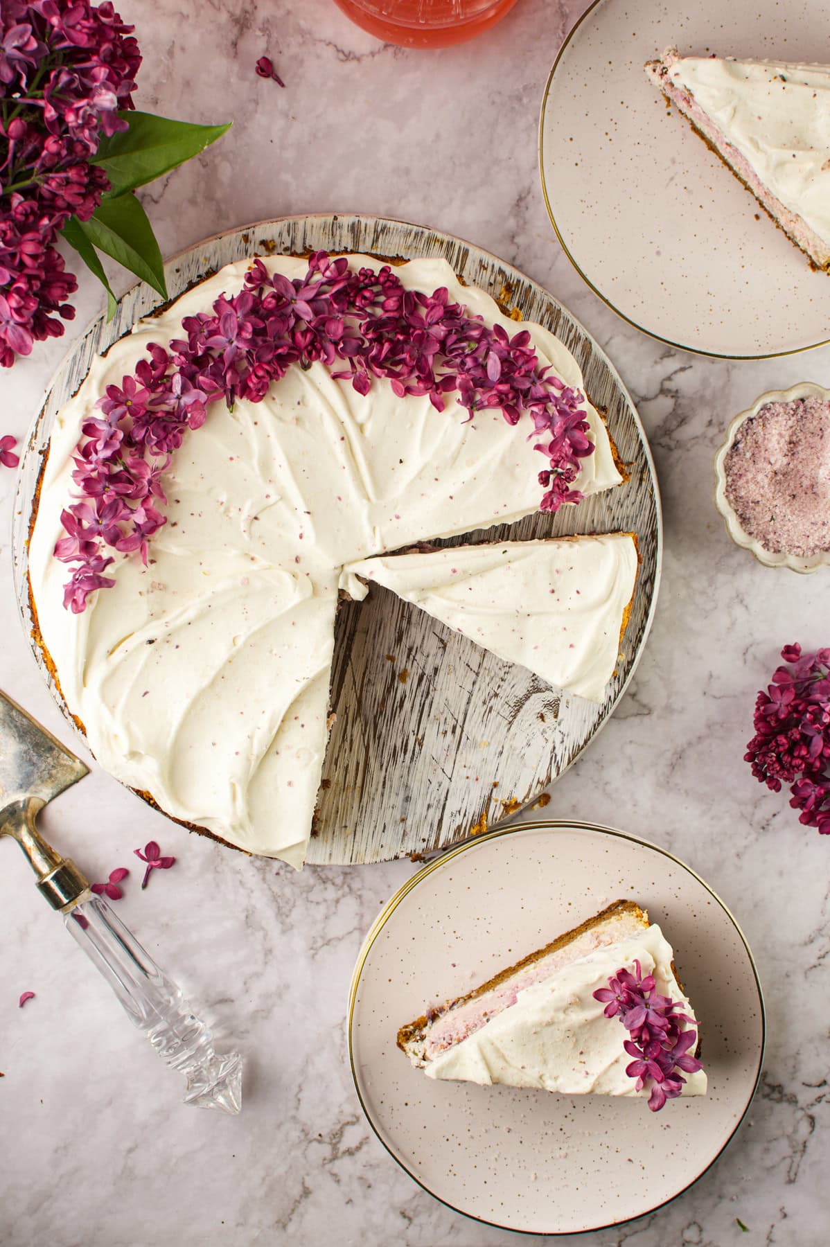 A Lilac Cheesecake with one slice cut and placed on a plate nearby. The cake sits on a marble surface, surrounded by lilac flowers and a small dish of purple powder.