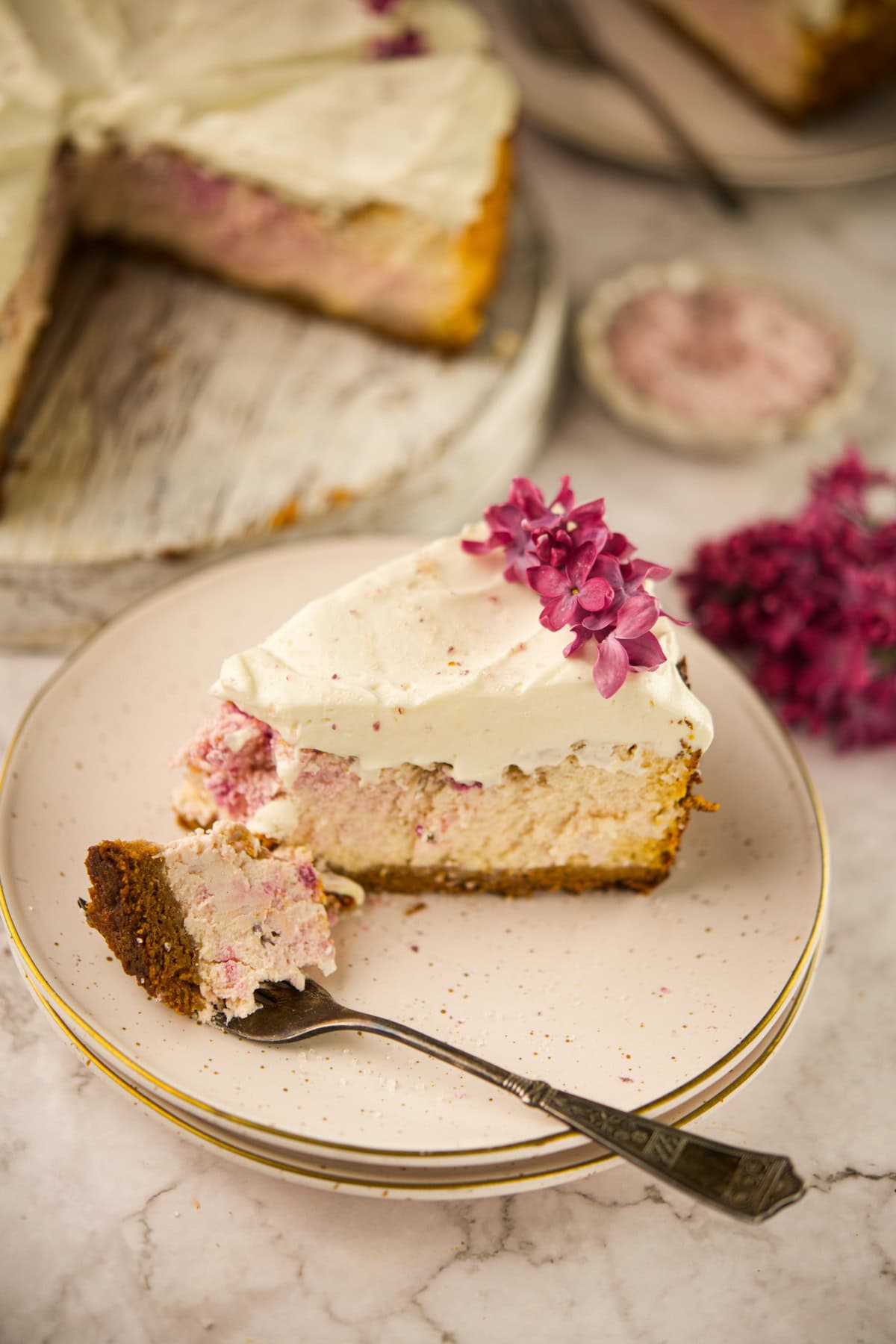 A slice of cheesecake with a creamy topping and pink flower garnish on a plate, with a fork holding a bite-sized piece; whole cheesecake and more flowers are visible in the background.