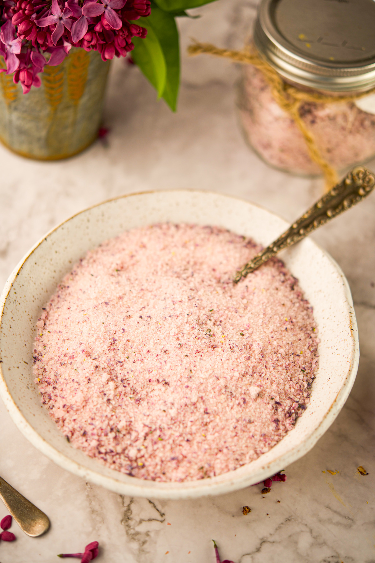 A white speckled bowl filled with pink lilac sugar sits on a marble surface, with a vintage spoon inside. A jar of lilac sugar and a vase of purple lilac flowers are in the background.