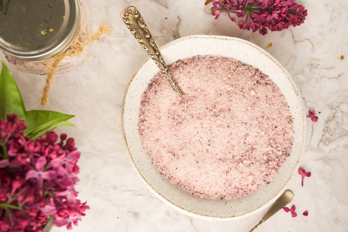 A bowl of pink and purple-hued sugar with a spoon, surrounded by fresh lilac flowers and a jar on a marble surface.