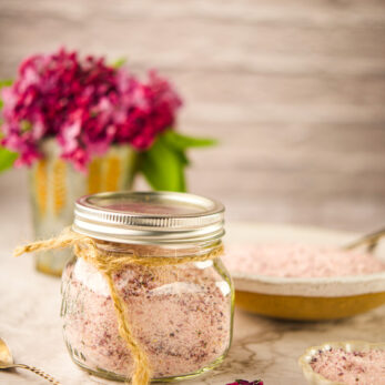 A glass jar filled with Lilac Sugar, tied with twine, sits on a marble surface. A bowl and small dish with more bath salts and a spoon are nearby. A vase of purple flowers is blurred in the background.