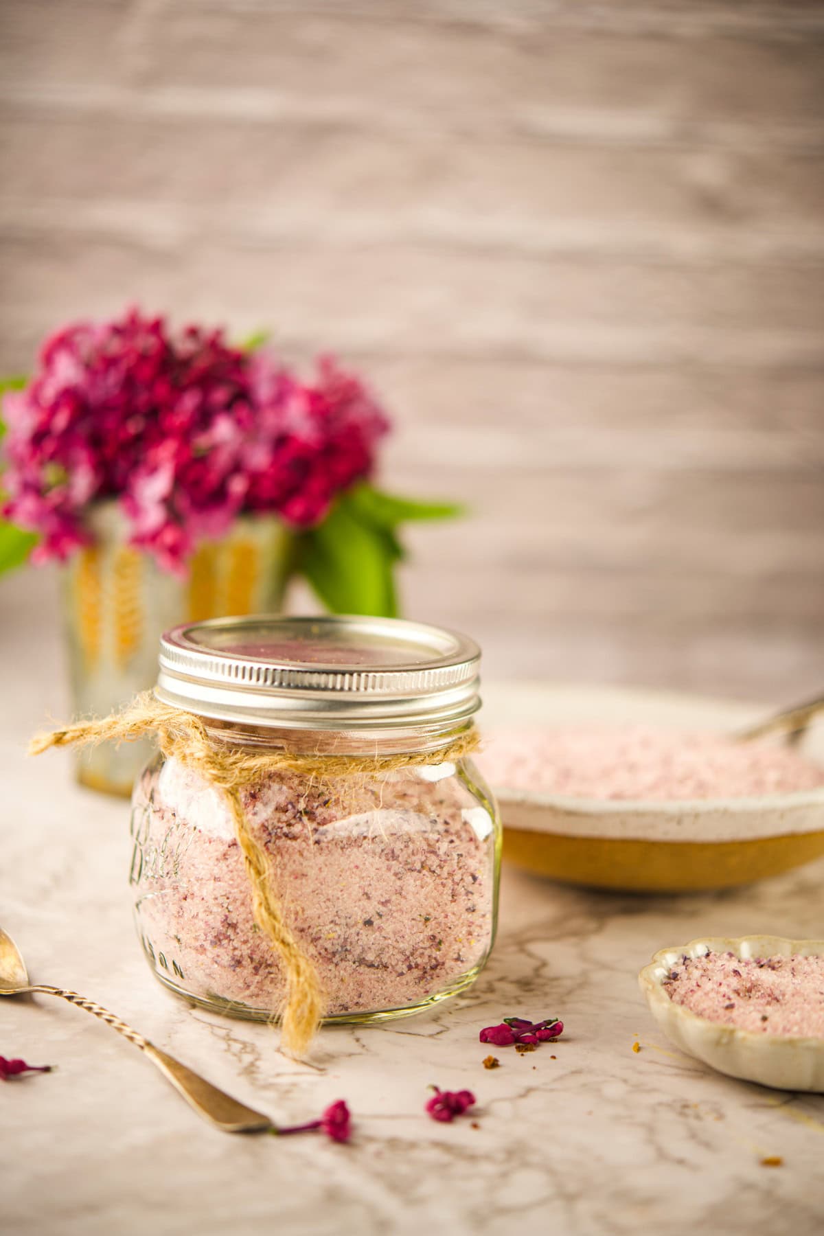 A glass jar filled with Lilac Sugar, tied with twine, sits on a marble surface. A bowl and small dish with more bath salts and a spoon are nearby. A vase of purple flowers is blurred in the background.