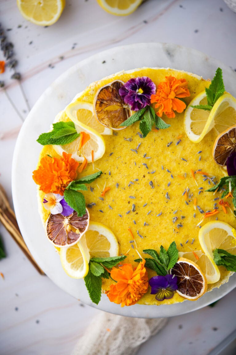 A round yellow cake decorated with lemon slices, dried orange slices, fresh mint leaves, edible flowers, and lavender, displayed on a white marble platter.