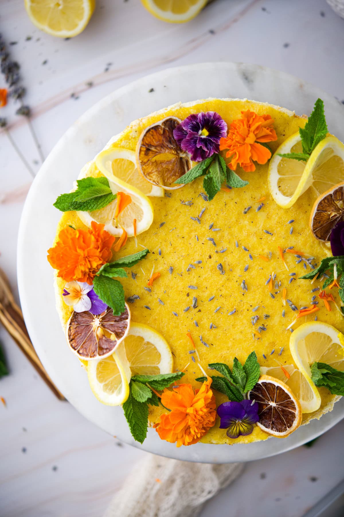 A round yellow cake decorated with lemon slices, dried orange slices, fresh mint leaves, edible flowers, and lavender, displayed on a white marble platter.