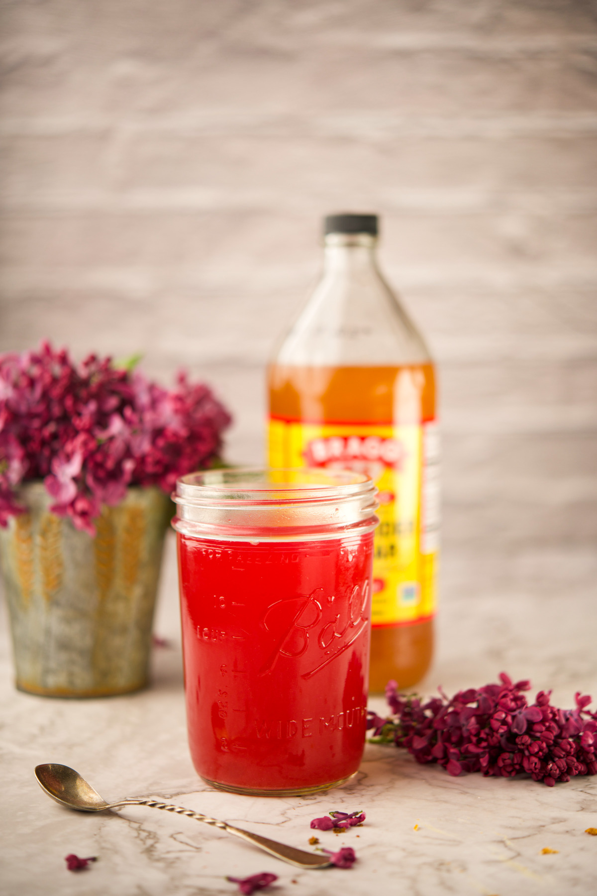 A mason jar filled with bright red liquid sits on a table next to a silver spoon, purple flowers, and a bottle of apple cider vinegar. A tin vase with more purple flowers is in the background.