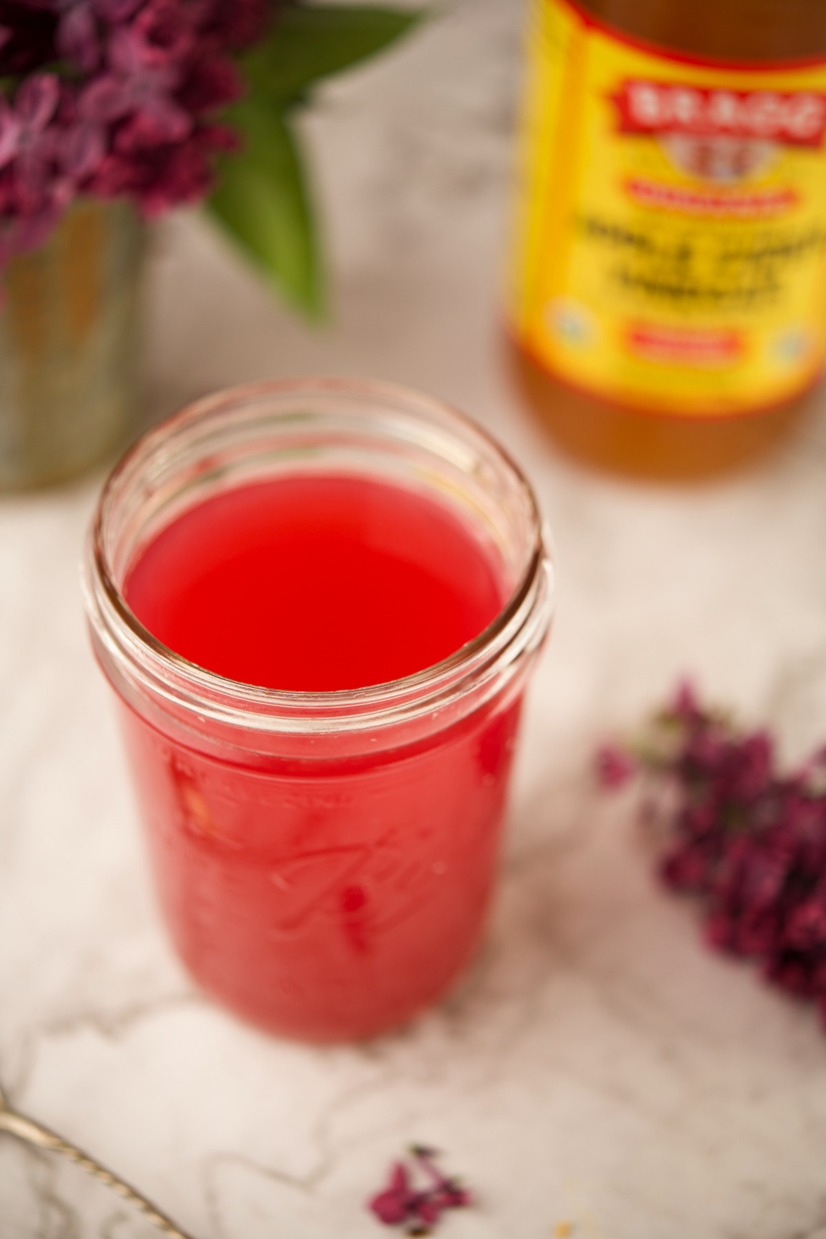 A glass jar filled with bright pink liquid sits on a marble surface, with a bottle of apple cider vinegar and purple flowers blurred in the background.