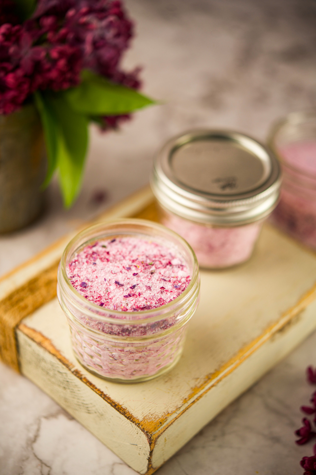 A small glass jar filled withLilac Flower Salt sits on a rustic wooden board, with additional jars and purple flowers blurred in the background.