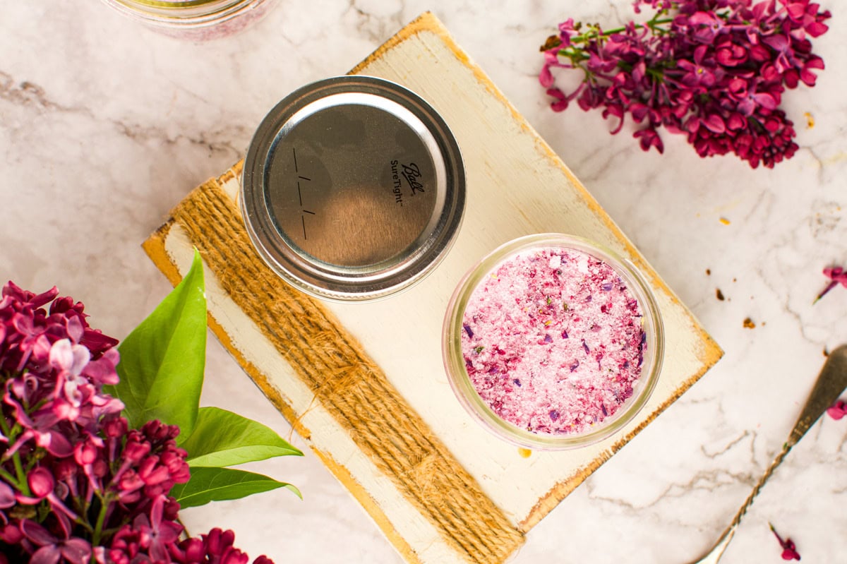 A jar of pink lilac sugar sits open on a wooden board, surrounded by fresh lilac flowers and leaves on a marble surface. The jar lid rests beside it.