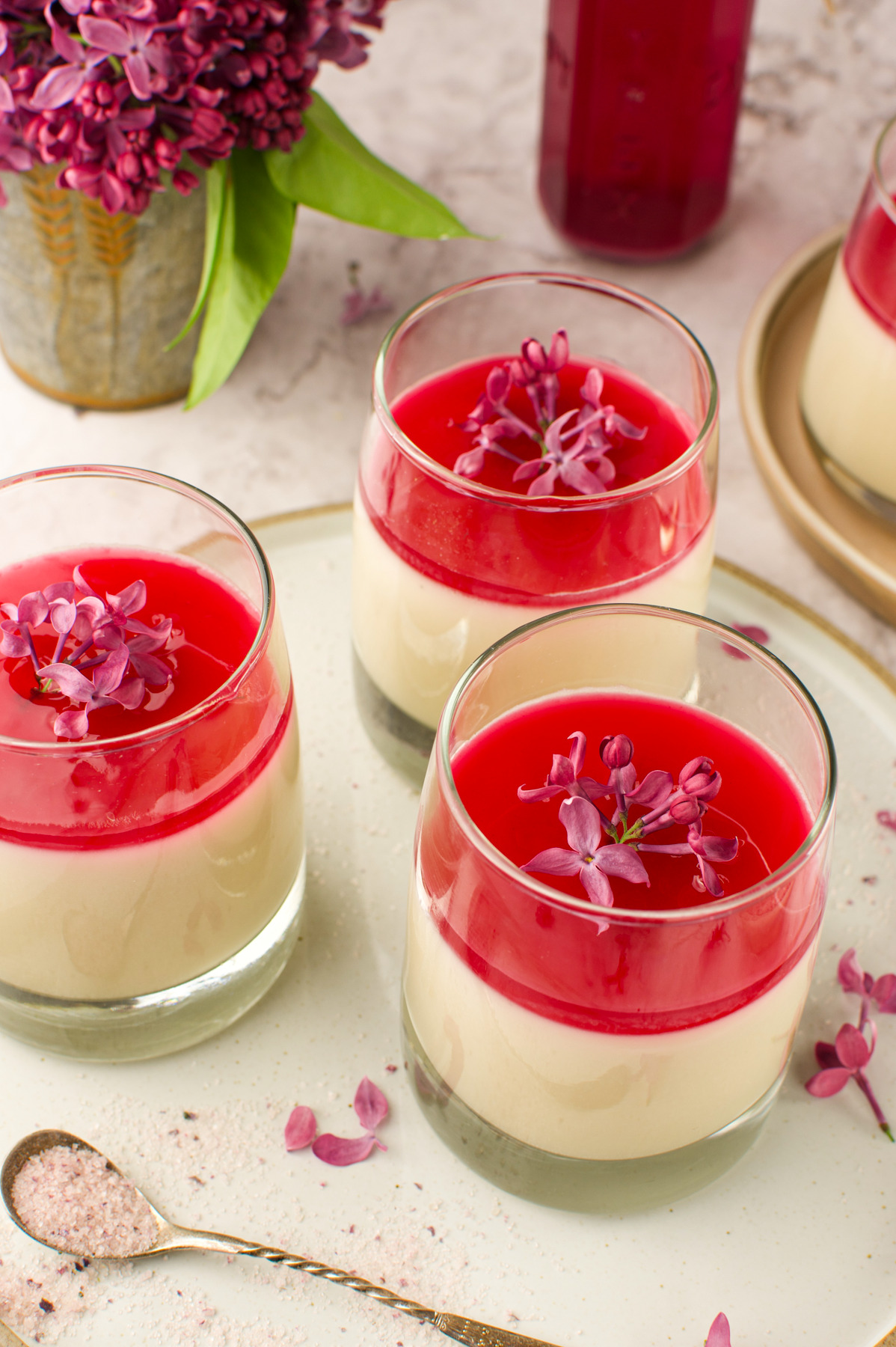 Three glasses of layered Coconut Panna Cotta With Lilac Syrup, each garnished with small purple flowers, are arranged on a white plate beside a spoon and a vase of lilacs.