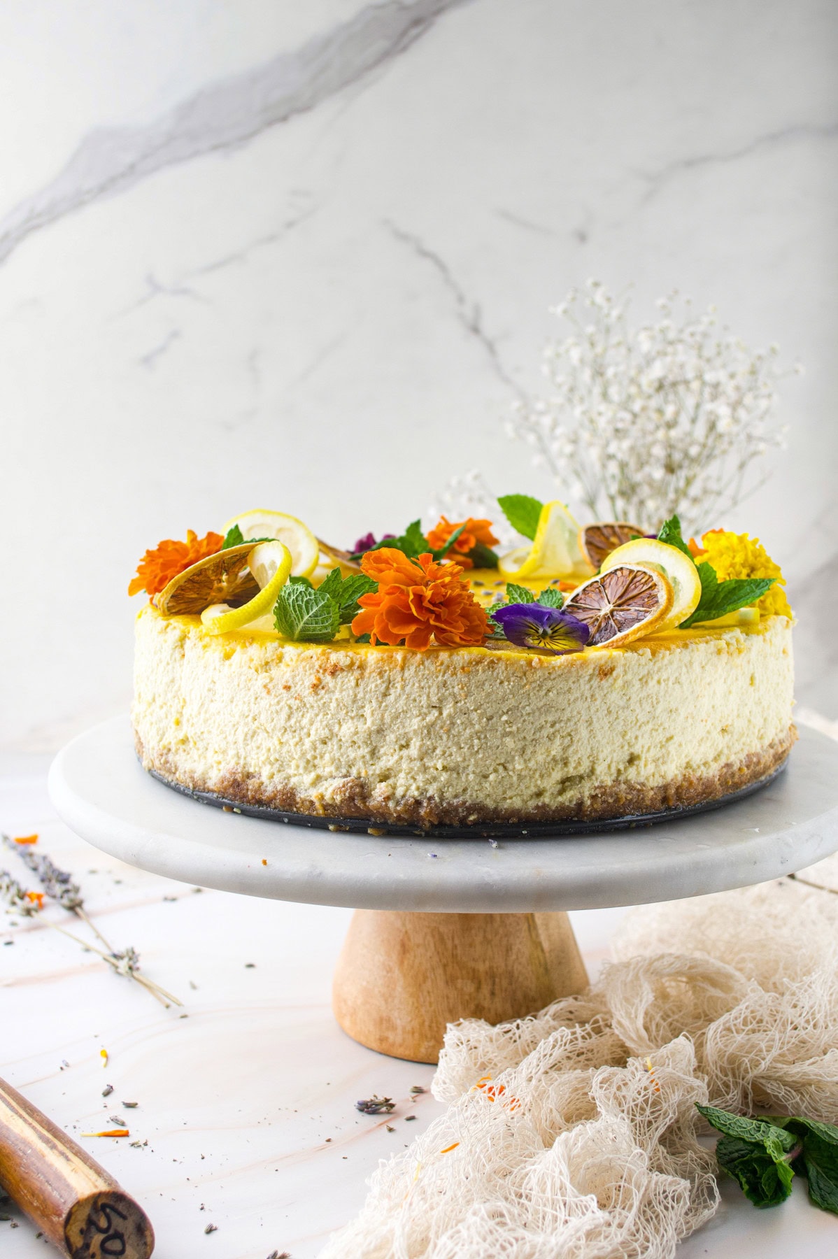 A cheesecake decorated with lemon slices, edible flowers, and fresh mint leaves sits on a marble cake stand against a white marble background, with delicate white flowers in the background.