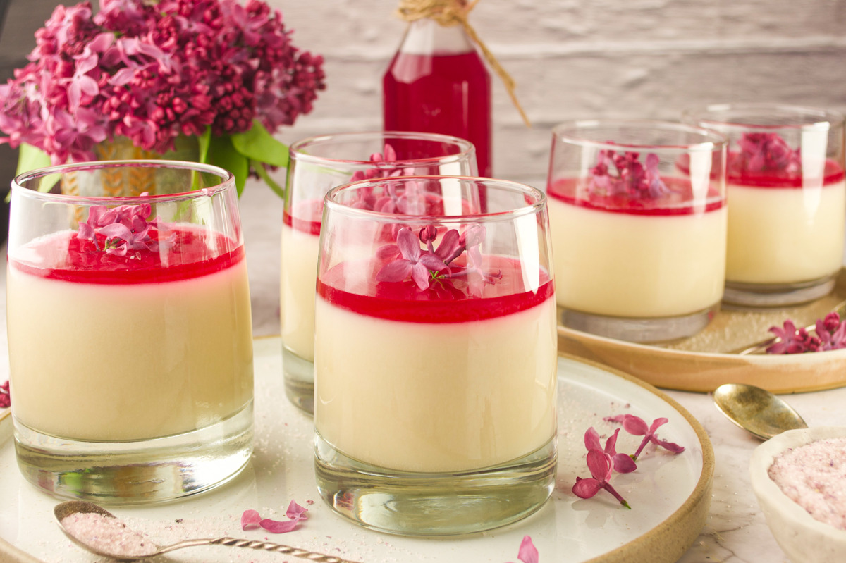 Five glasses of creamy dessert topped with a bright pink layer and garnished with purple flowers are arranged on a tray, with lilac blossoms and a bottle of pink syrup in the background. Spoons and a bowl are also visible.