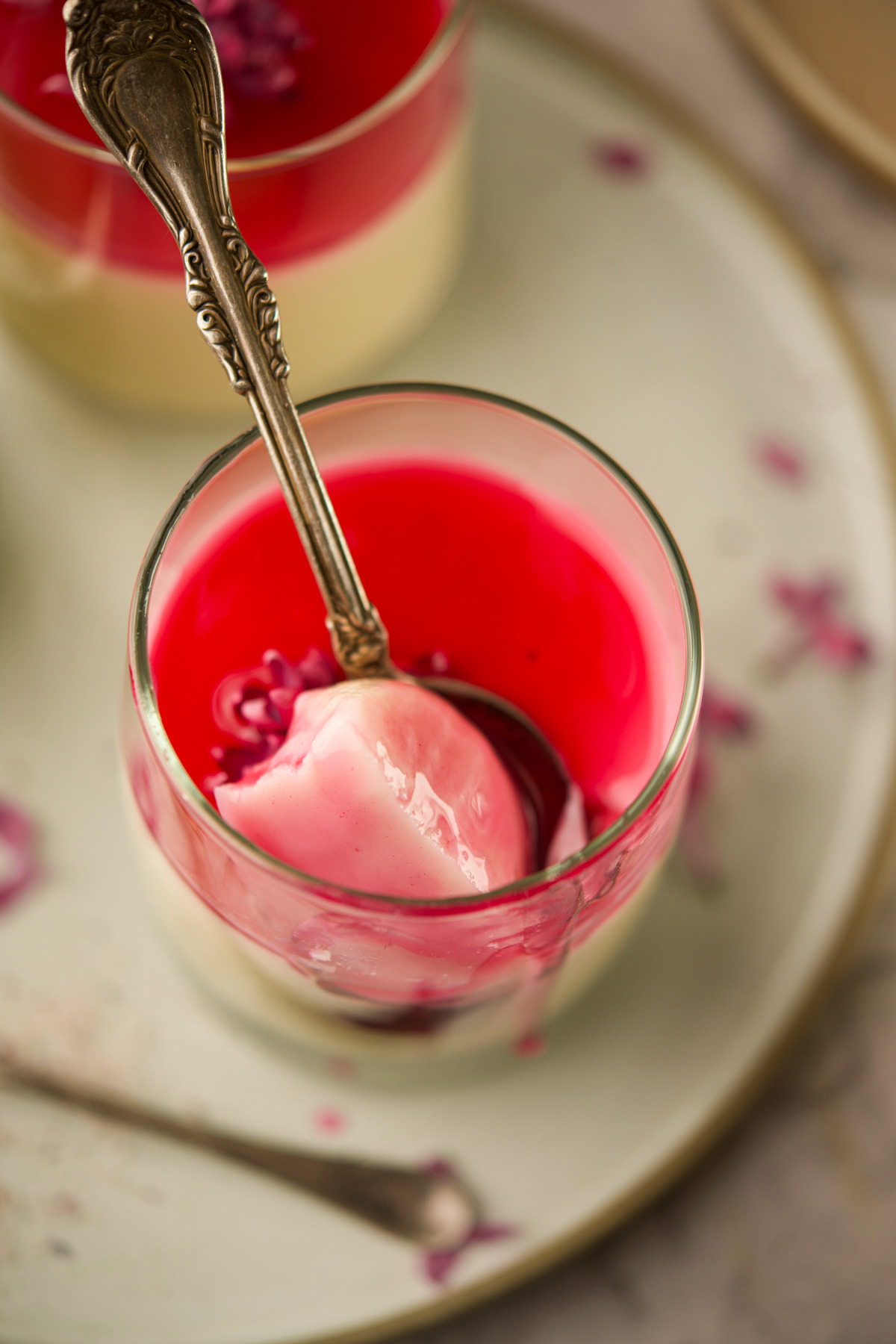 A glass dessert cup filled with layered panna cotta; the top layer is bright red jelly. A spoon rests inside, scooping some panna cotta. Another cup and spoon appear blurred in the background.