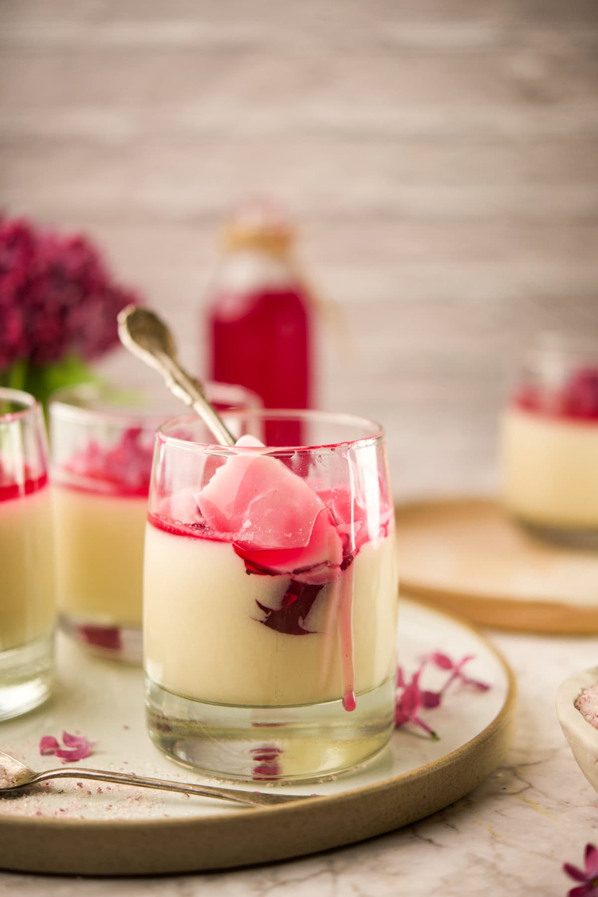 A glass of creamy white dessert topped with a layer of pink jelly and sauce, served with a spoon. More glasses with similar desserts and a bottle of pink liquid are in the background on a decorative tray.