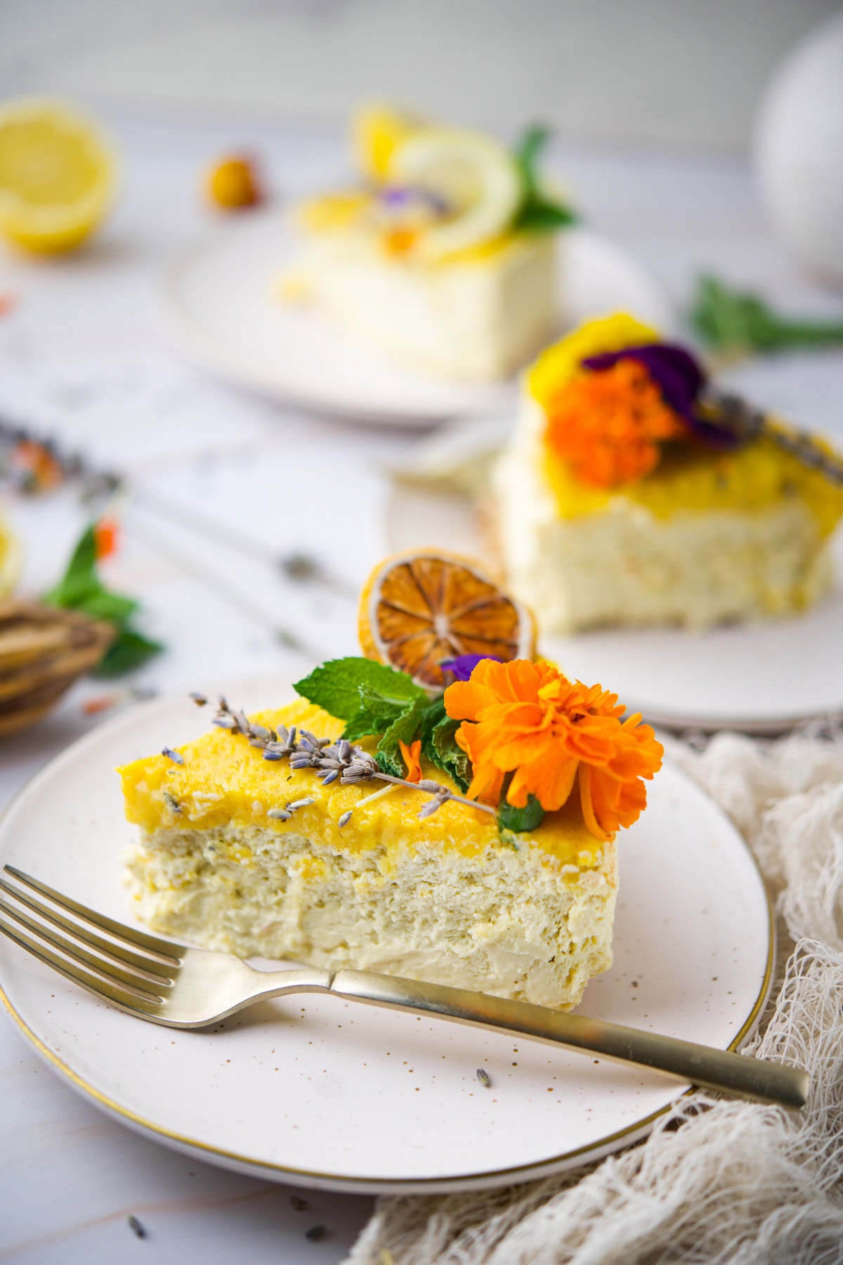 A slice of cake topped with yellow icing, edible flowers, dried citrus, herbs, and lavender, served on a white plate with a gold fork. Two more decorated cake slices are blurred in the background.