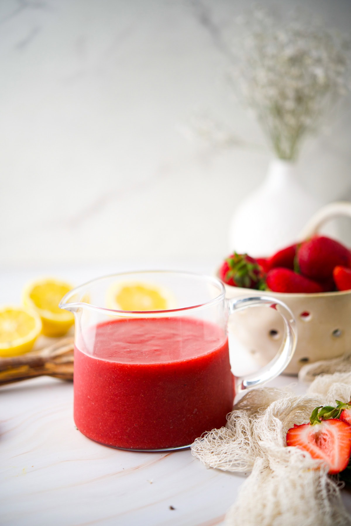 A glass pitcher filled with bright red strawberry sauce sits on a white surface, surrounded by fresh strawberries, lemon halves, and a white colander. Blurred flowers and fruit are visible in the background.