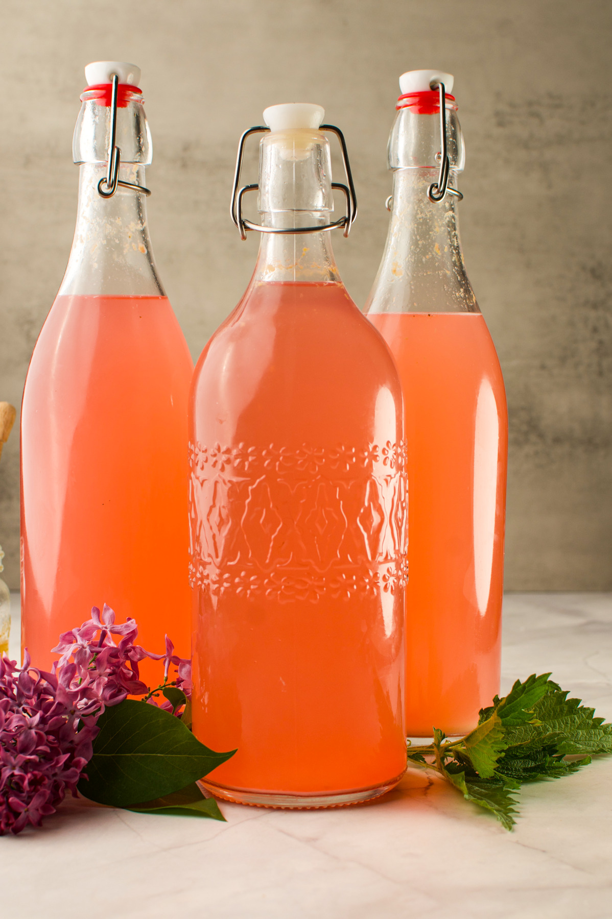 Three glass bottles filled with pink liquid are displayed on a light surface, with lilac flowers and green leaves arranged nearby. The bottles have swing-top caps and decorative patterns.