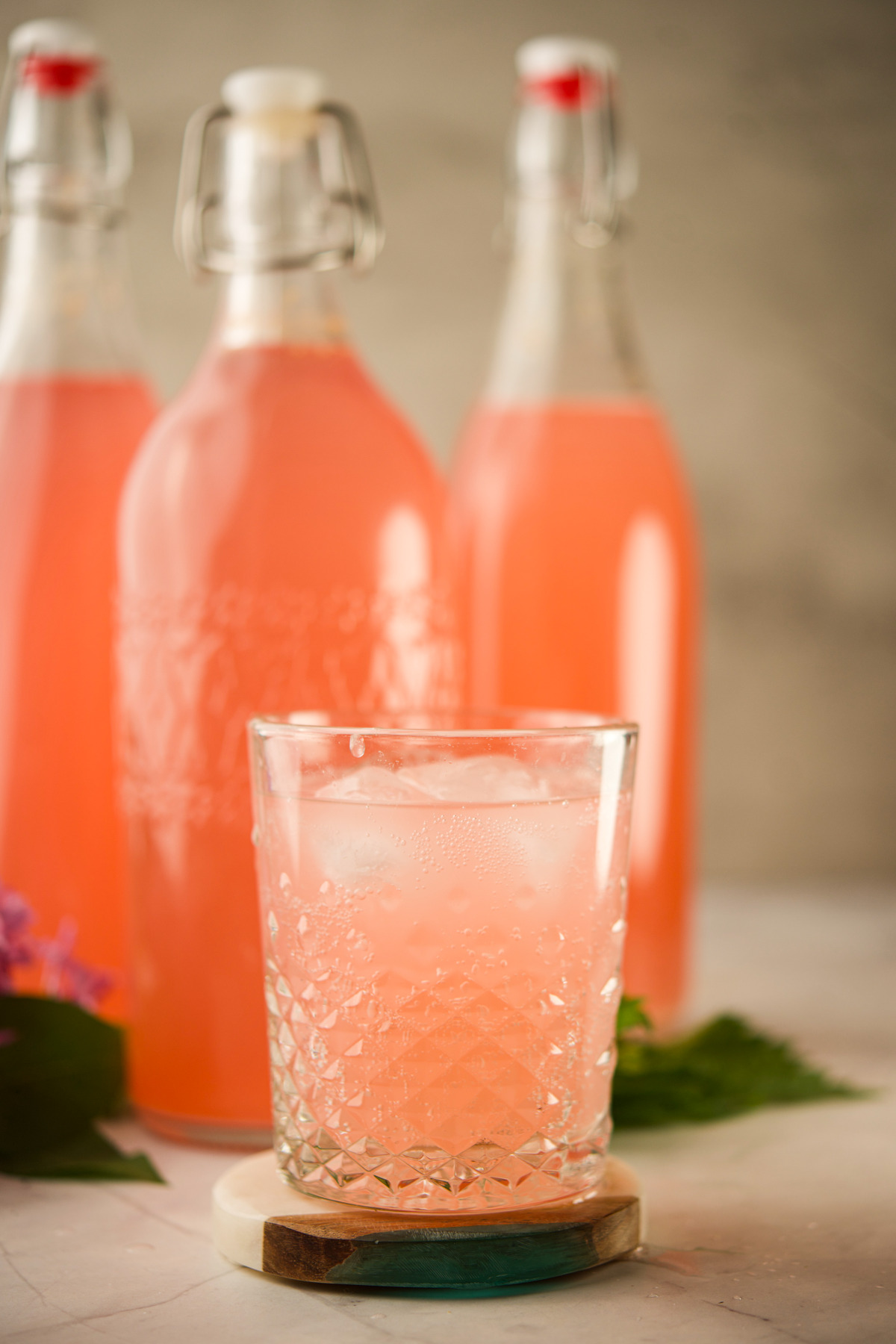 A glass of iced pink drink sits in front of two tall bottles filled with the same pink beverage, with a few leaves and purple flowers placed nearby on a light surface.