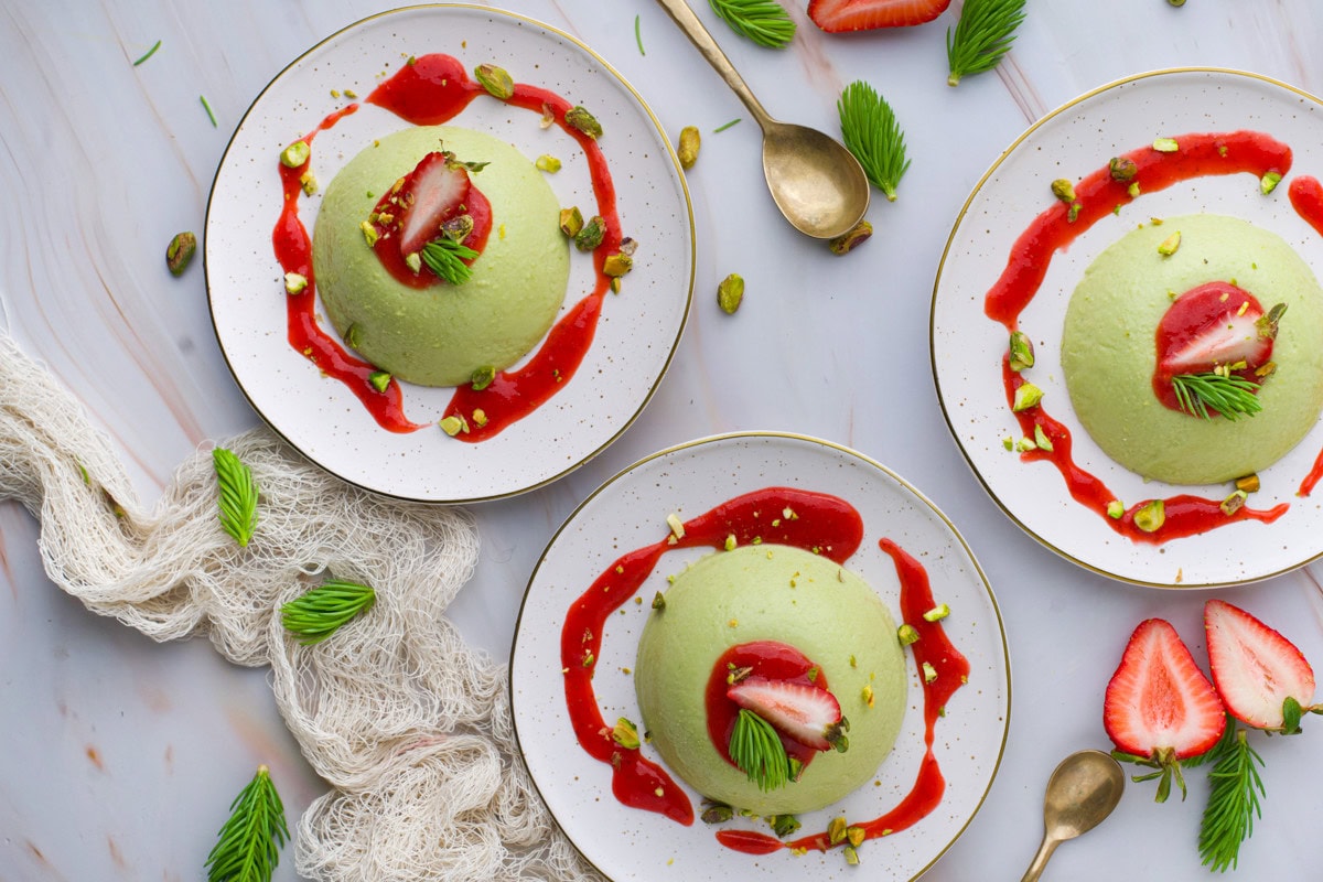 Three plates with green mousse desserts, each topped with a sliced strawberry and green garnish, surrounded by red sauce and chopped pistachios. Golden spoons, strawberries, and herbs are scattered on a marble surface.