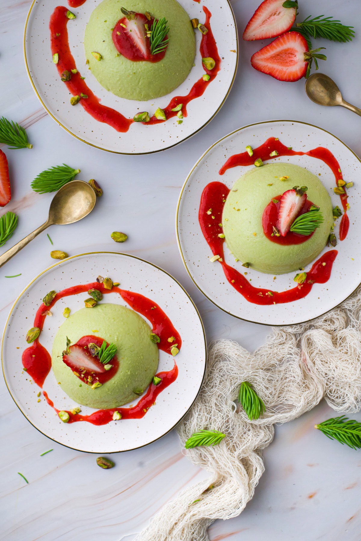 Three plates with dome-shaped green desserts topped with strawberry slices, fresh green leaves, and chopped pistachios, each surrounded by a swirl of red sauce, with gold spoons and strawberries on a marble surface.