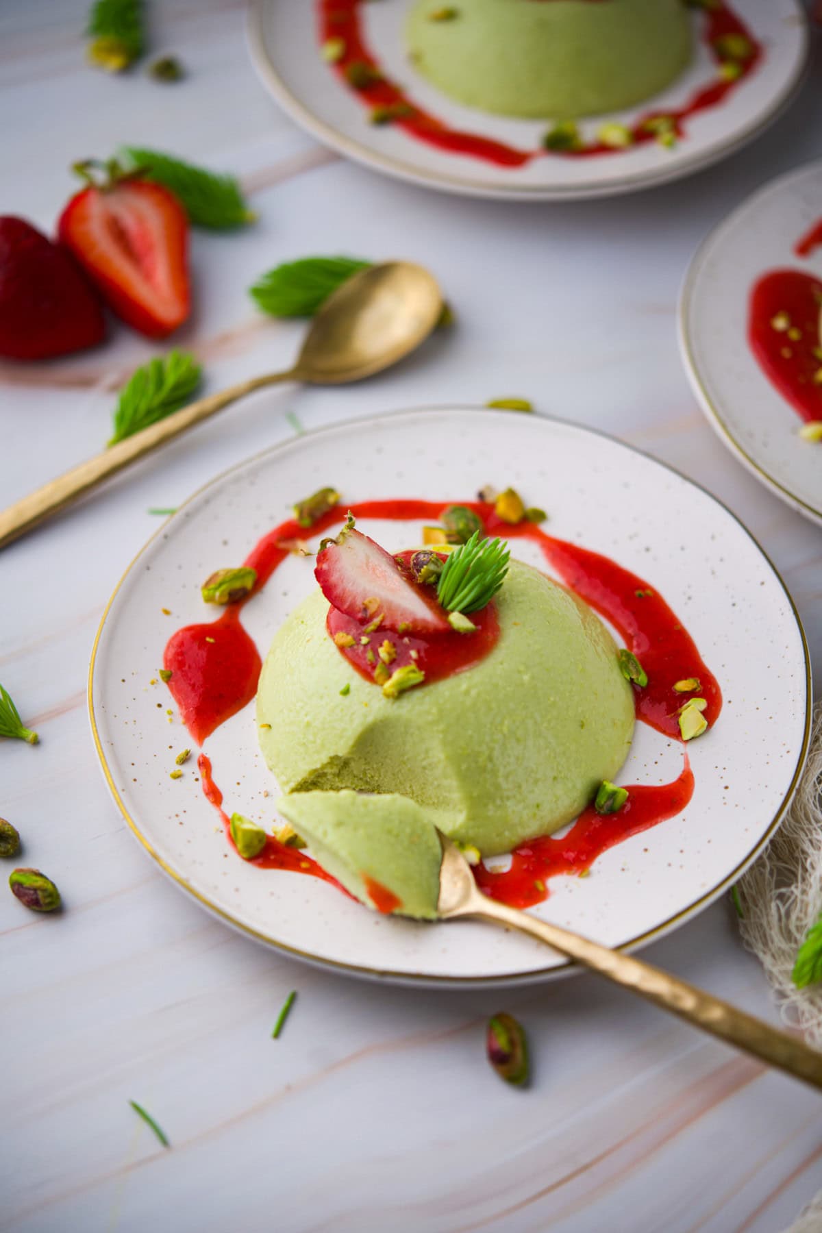 A dome-shaped green dessert topped with a sliced strawberry, chopped pistachios, and mint. Strawberry sauce is drizzled around it on a white plate with a gold spoon, with additional plates and strawberries in the background.