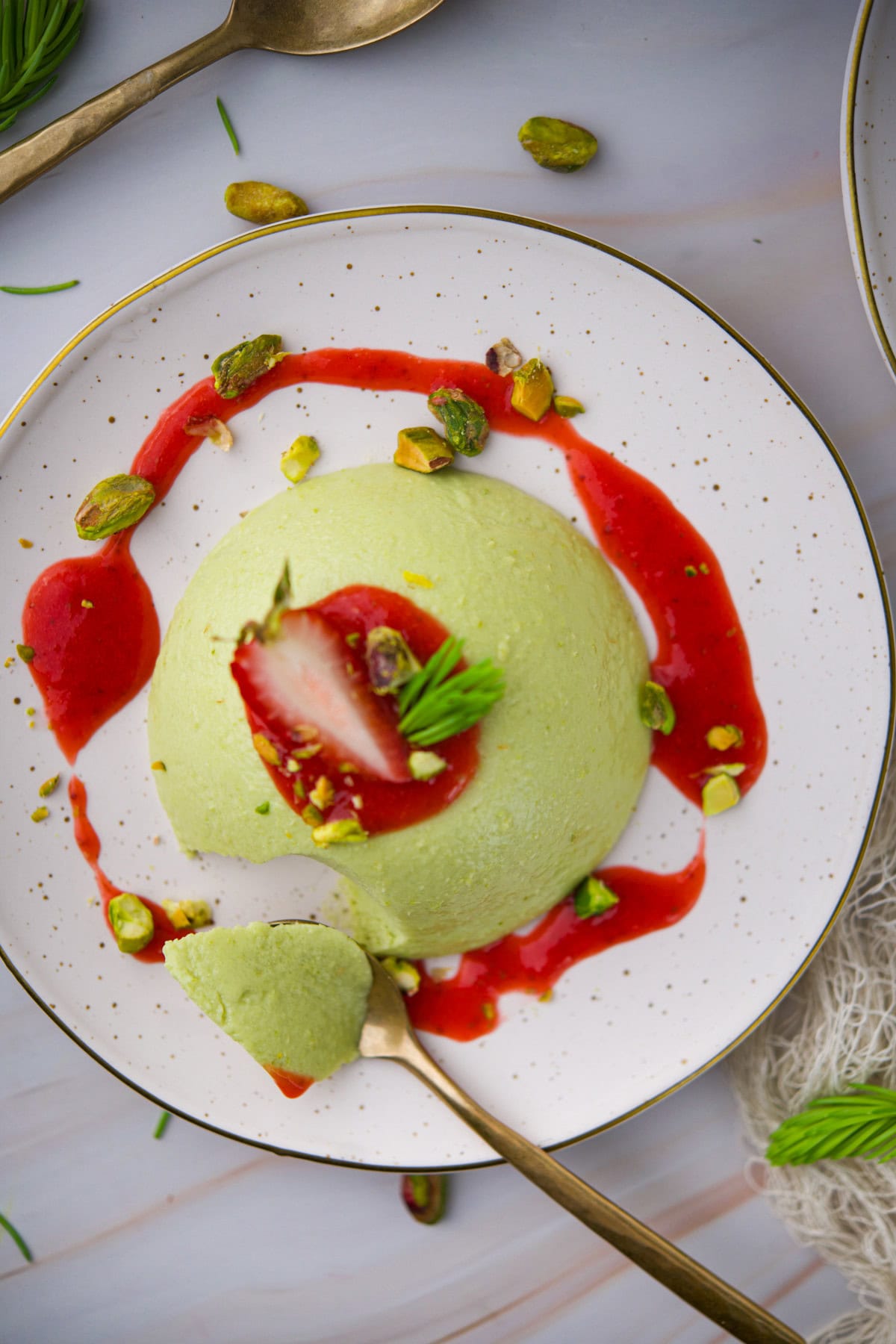 A creamy Coconut Spruce Tip Panna Cotta topped with a strawberry slice, pistachios, and a bright red sauce. A gold spoon takes a bite from the dessert on a white plate with a gold rim, garnished with herbs.