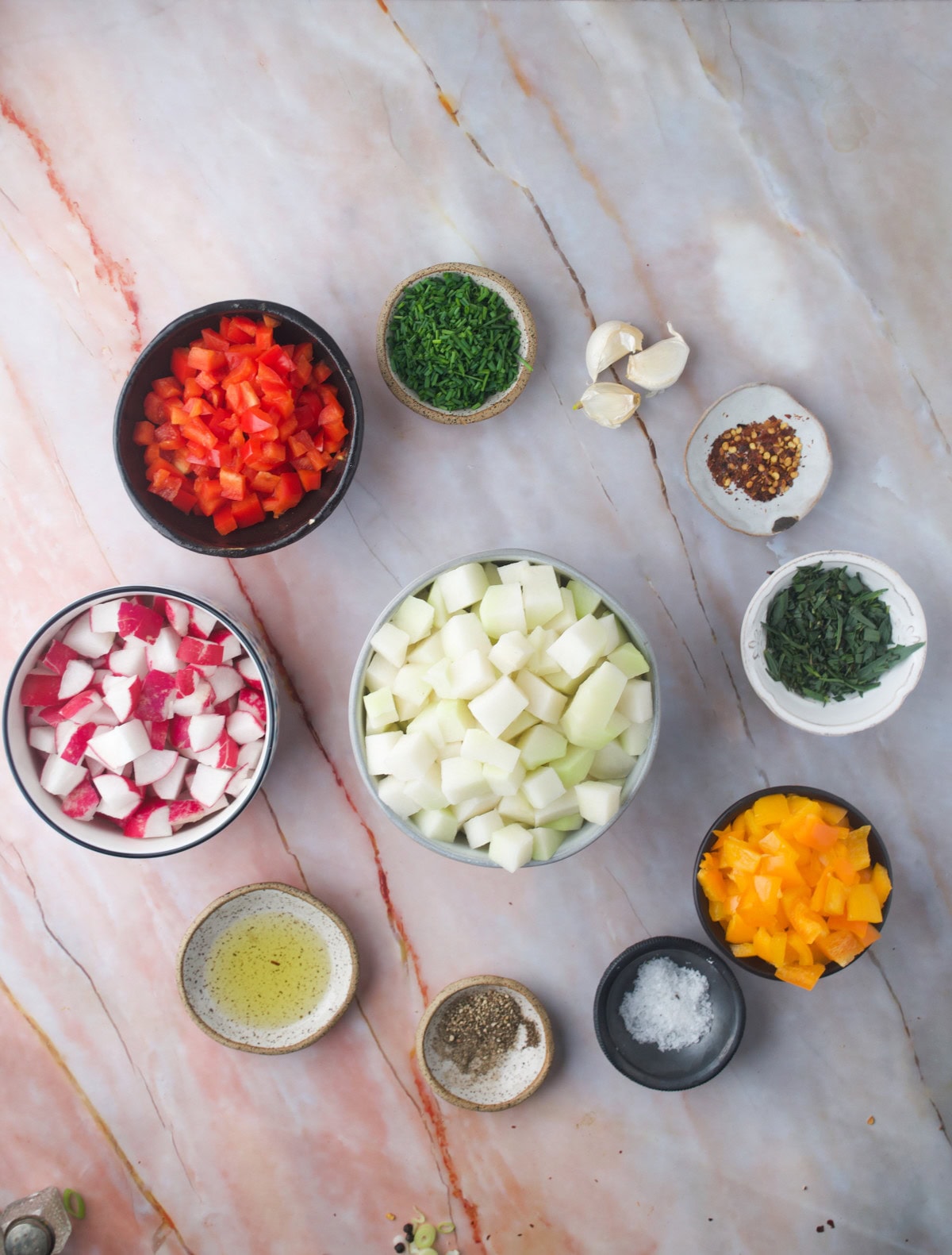 Bowls containing diced red and yellow bell peppers, chopped onion, diced radish, fresh herbs, garlic cloves, olive oil, salt, pepper, and chili flakes on a pink marble surface.