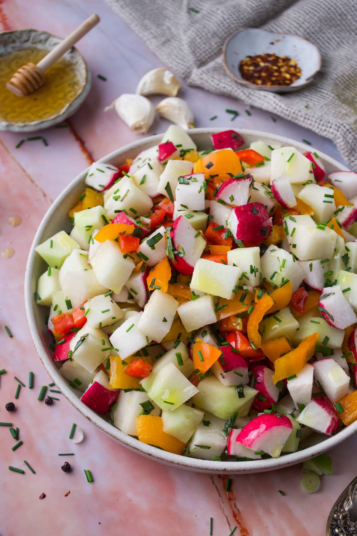 A bowl of chopped vegetable salad with radishes, bell peppers, and chives sits on a pink marble surface, surrounded by a honey dipper, garlic cloves, and a small dish of chili flakes.