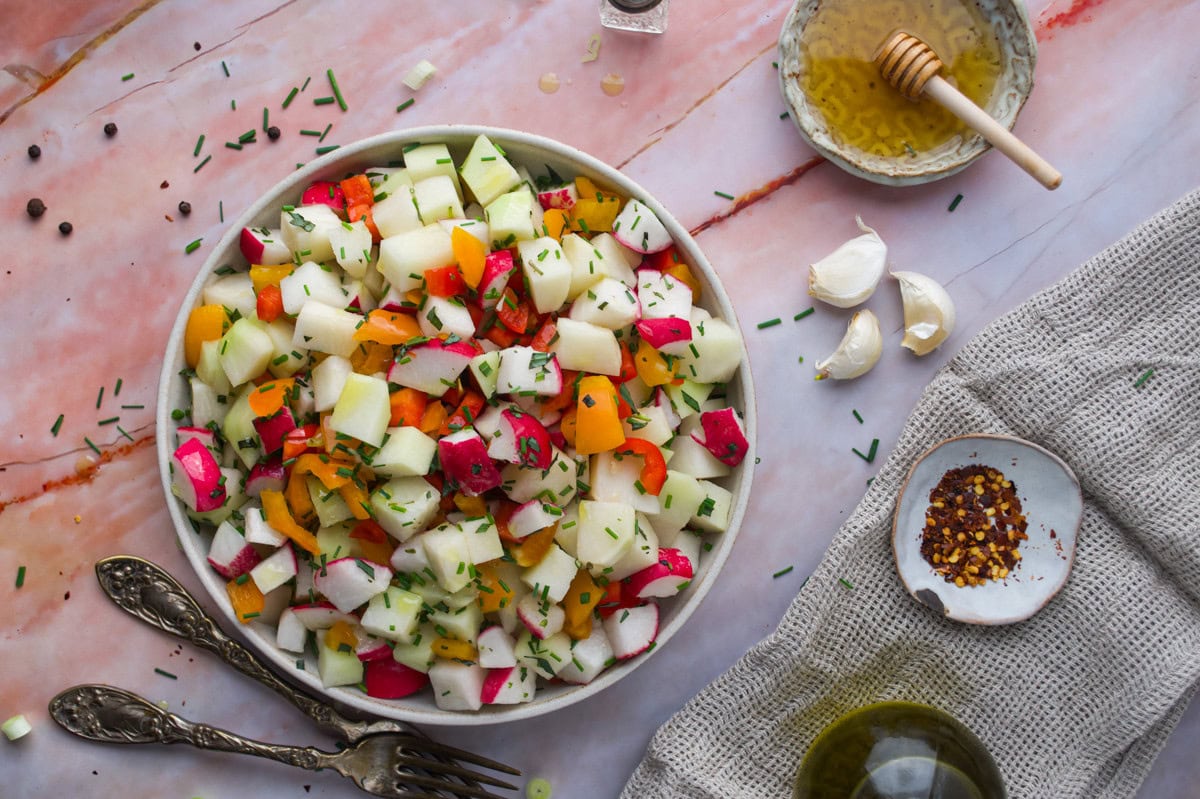 A bowl of colorful Chopped Kohlrabi Salad salad with radish, cucumber, and yellow peppers sits on a pink marble surface, surrounded by garlic, chives, olive oil, honey, and chili flakes.