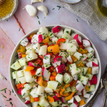 A bowl of colorful Chopped Kohlrabi Salad garnished with herbs. Garlic cloves, honey, red pepper flakes, olive oil, and utensils are nearby on a marble surface.