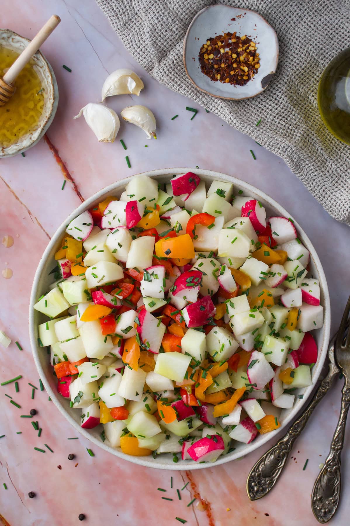 A bowl of colorful chopped salad with cucumbers, radishes, and bell peppers, garnished with herbs. Garlic cloves, honey, red pepper flakes, olive oil, and utensils are nearby on a marble surface.
