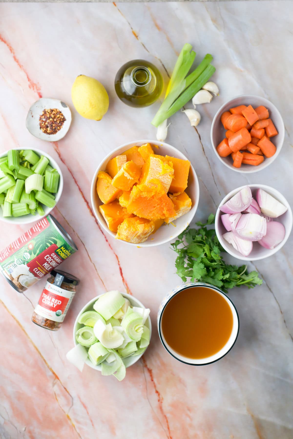 Ingredients for a soup laid out on a marble surface, including pumpkin chunks, carrots, celery, shallots, leeks, garlic, spring onions, cilantro, vegetable broth, olive oil, lemon, red pepper flakes, tomato paste, and canned tomatoes.